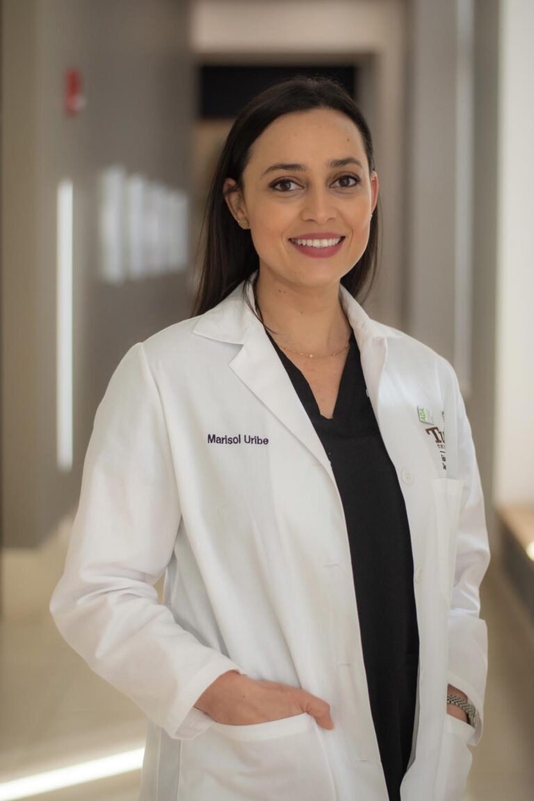 Dr. Marisol Uribe wearing a white lab coat embroidered with the name “Marisol Uribe,” standing in a blurred indoor hallway; black shirt visible underneath.