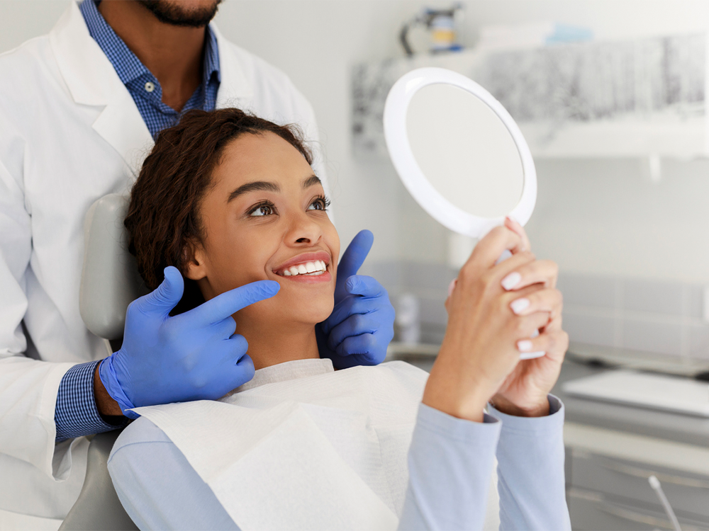 A woman smiling while holding a mirror with a dentist beside her.